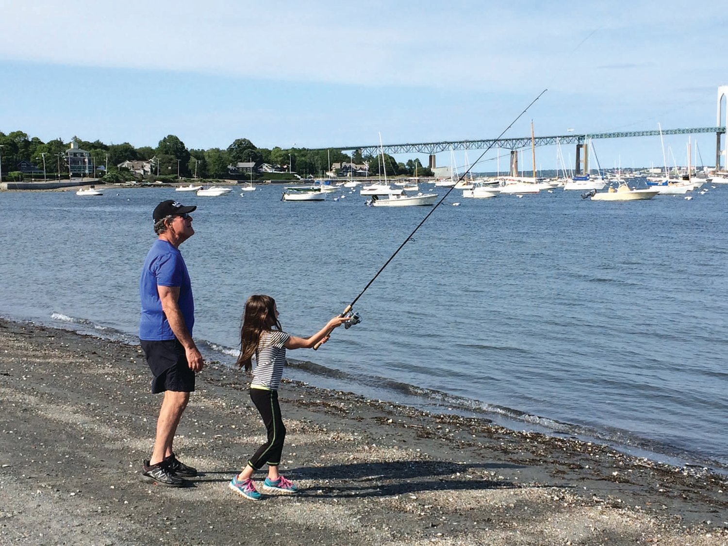 PRACTICING YOUR CAST: Capt. Ken Cooper with his granddaughter Rayna (then 7 years old) practicing casting in Jamestown with the Newport
Bridge in the background.